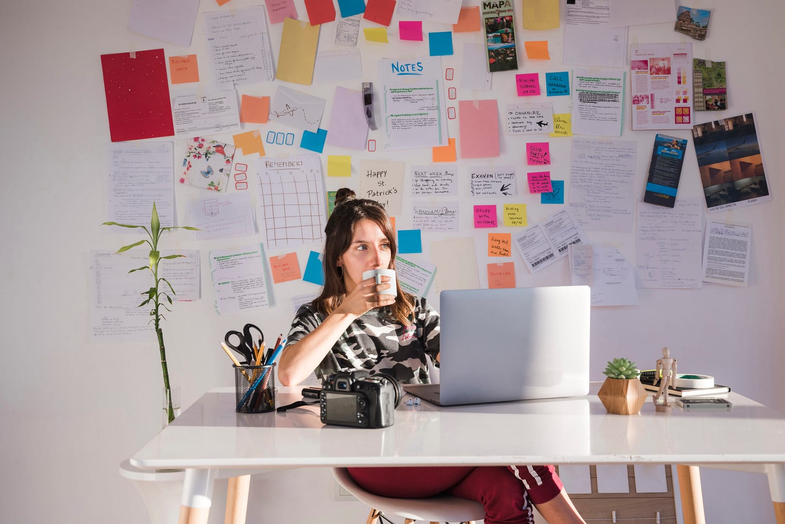 A female freelancer sipping coffee while working at a laptop, surrounded by colorful notes and ideas pinned on the wall