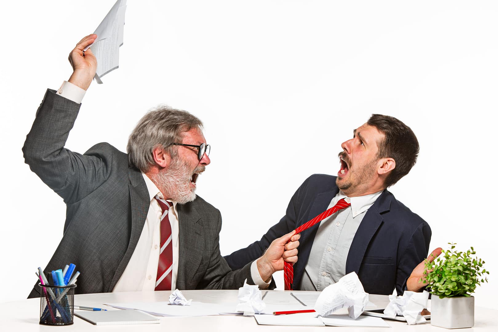 An angry older man in a suit holding a document, scolding a younger man while pulling his tie in frustration. The younger man appears shocked and defensive, seated at a desk cluttered with crumpled paper, pens, and documents