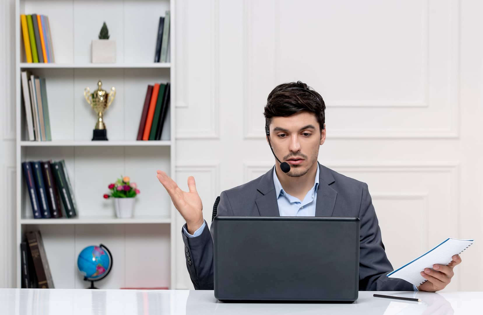 Man in a grey suit with a headset holding a notepad, discussing during an online meeting, symbolizing visibility and control in workplace communication.